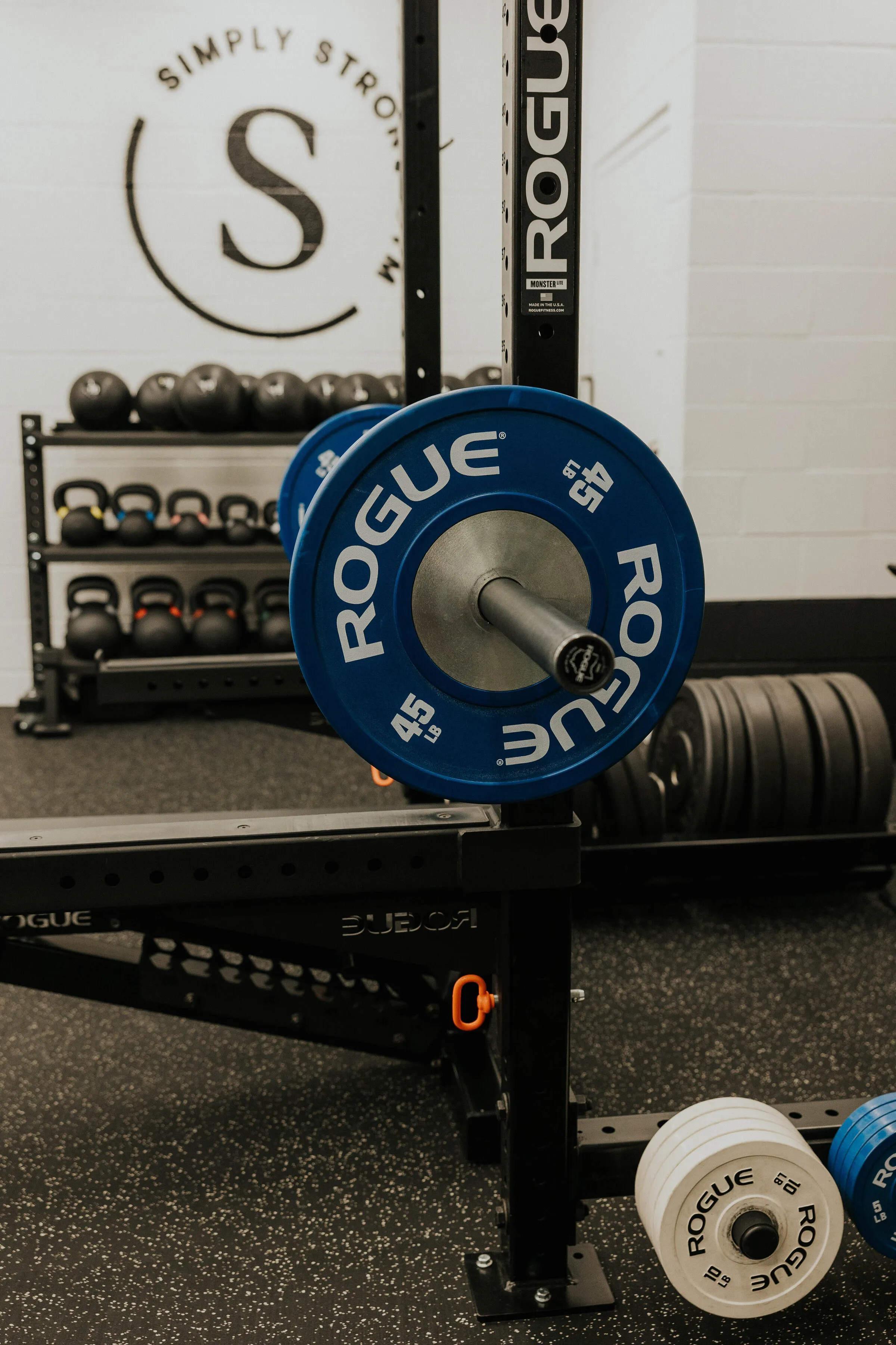 A barbell loaded with Rogue bumper plates in front of the Simply Strong Gym logo